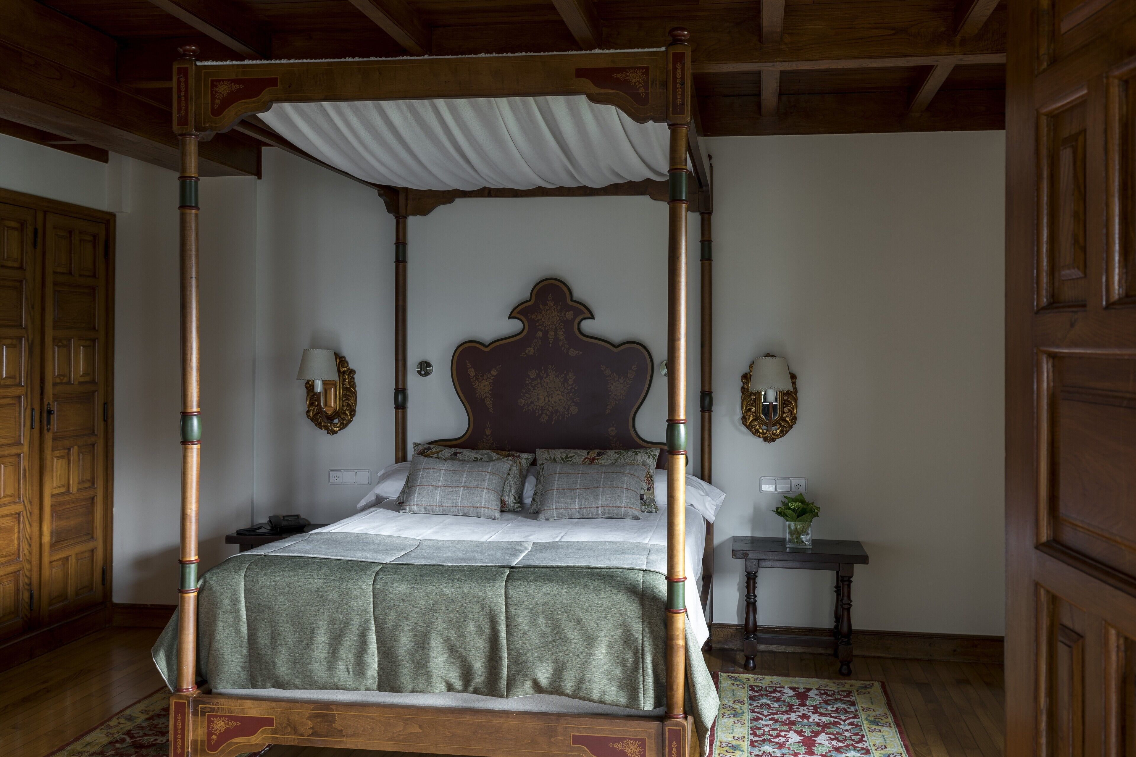 A four-poster canopy bed with a wooden frame and a white canopy, flanked by two wall sconces and a bedside table with a vase of flowers.
