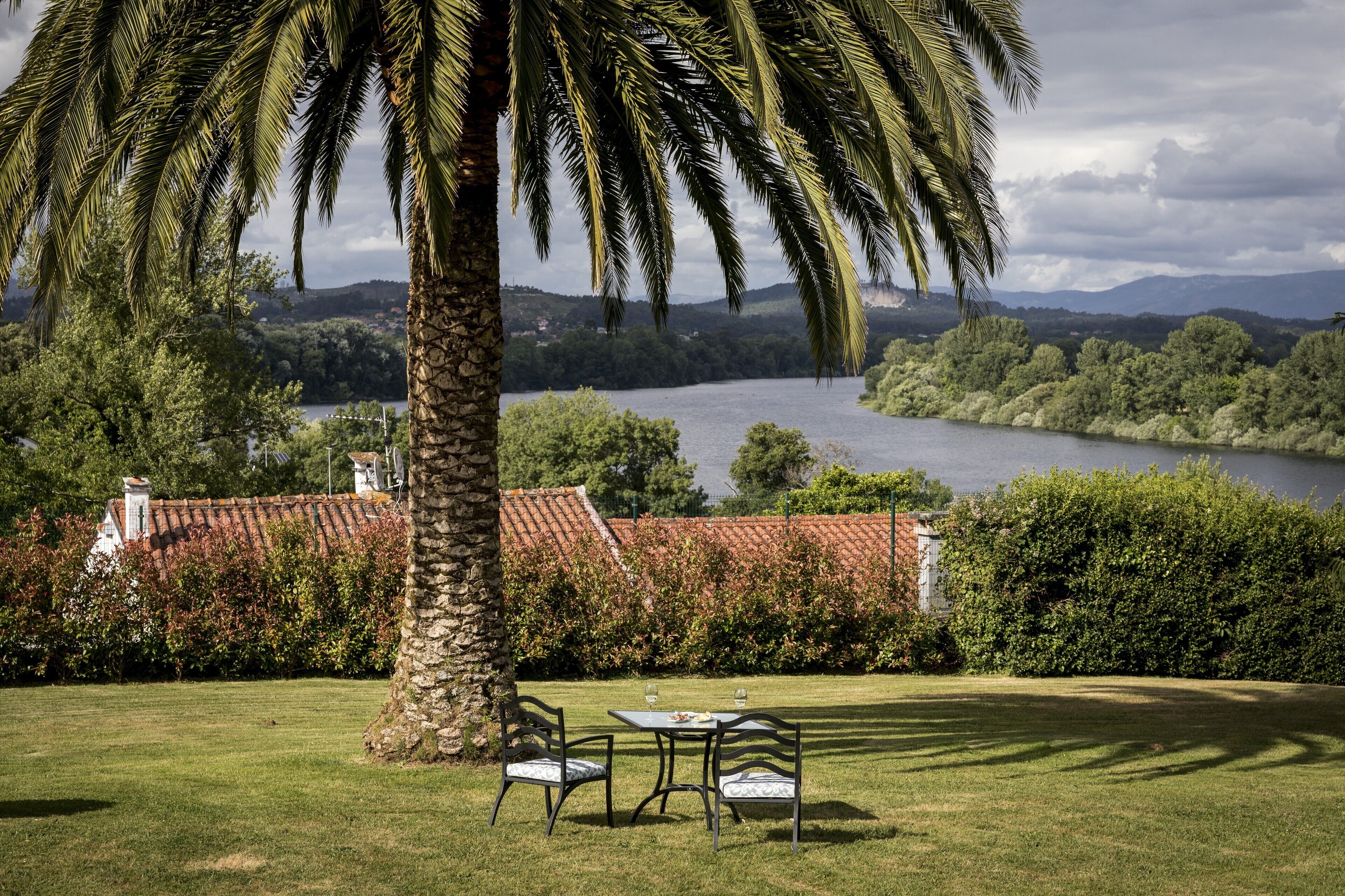 Un espacio al aire libre con césped, dos sillas y una mesa, con vistas a un lago y colinas a lo lejos.