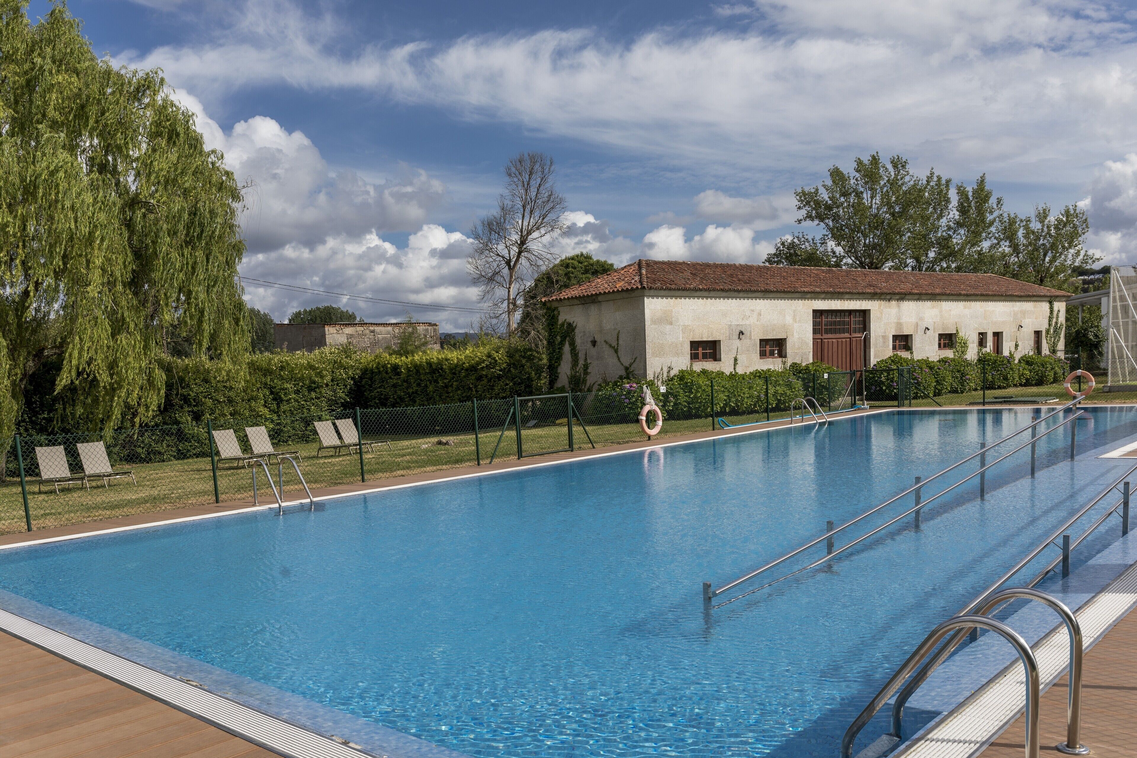 A swimming pool with a wooden deck, a building with a tiled roof, and a fence with lounge chairs.