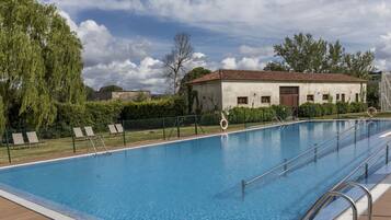 A swimming pool with a wooden deck, a building with a tiled roof, and a fence with lounge chairs.