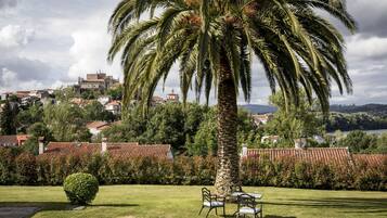 Un jardin avec un palmier, une petite table entourée de chaises, et une vue sur une ville où l’on aperçoit, au loin, des bâtiments aux toits rouges.