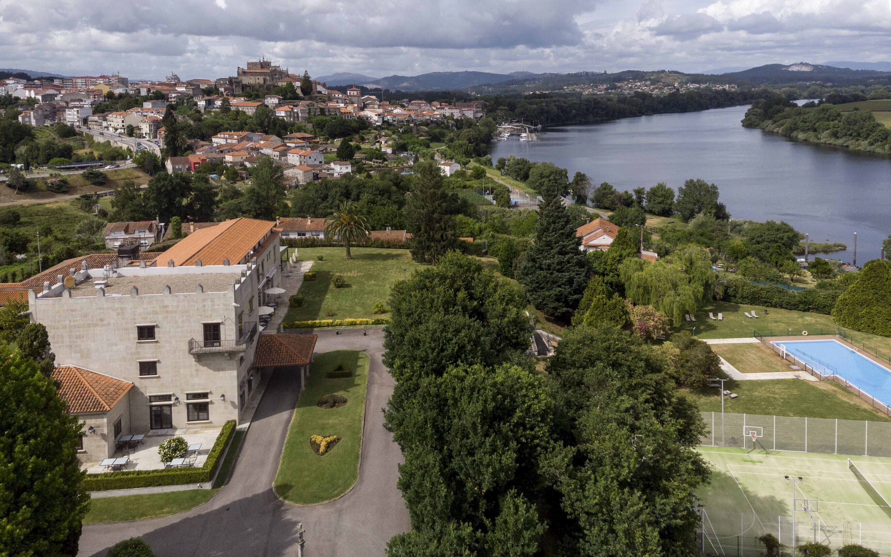 An aerial view of a town with a prominent castle-like structure, a river, and a well-maintained park with a swimming pool.