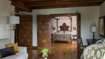 A spacious room with a wooden ceiling, a bed with a wooden headboard, a sofa, a coffee table with a magazine, and a door leading to another room.