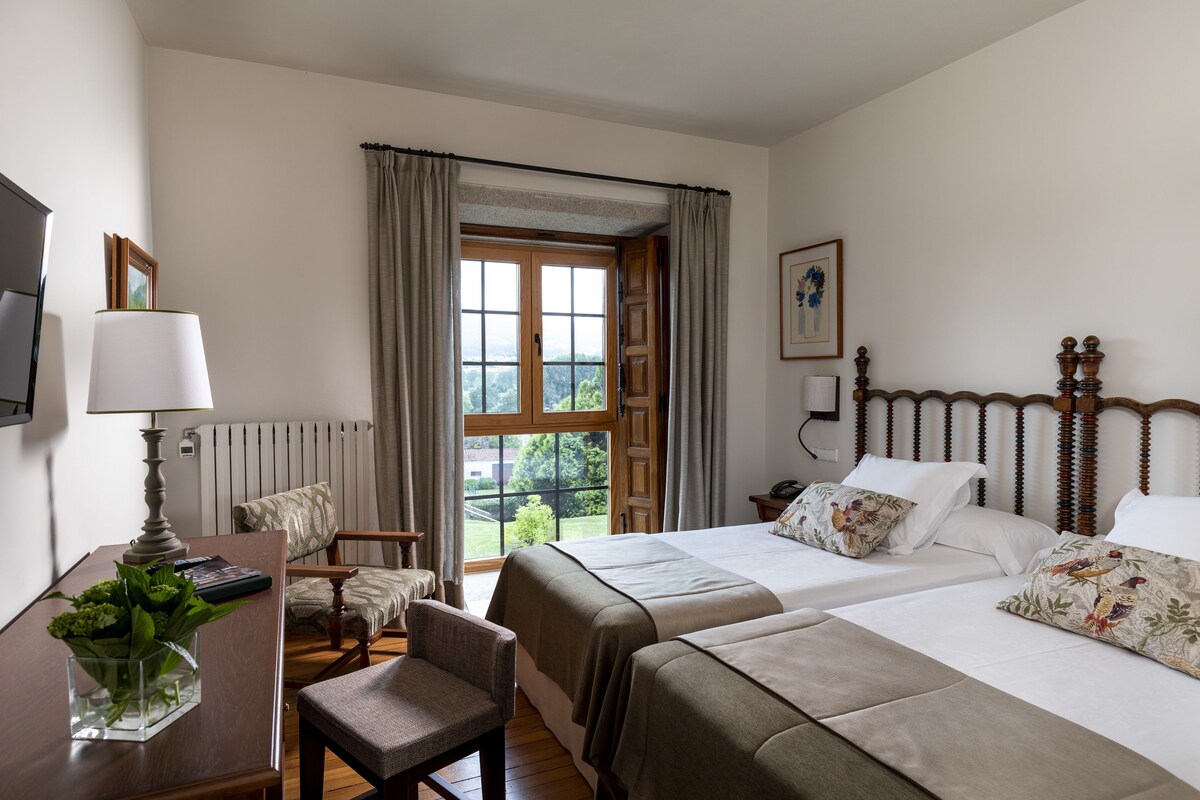 A hotel room with two beds, a desk with a lamp, a chair, and a view of the outdoors.