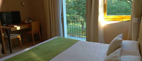 Habitación de hotel con cama, escritorio, silla, televisión y balcón con vistas a árboles.