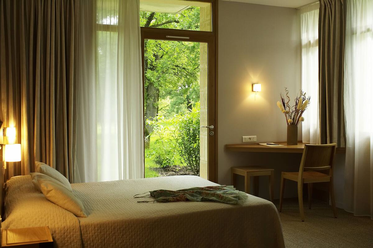 A hotel room with a bed, a desk, and a view of greenery through a door.