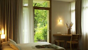 A hotel room with a bed, a desk, and a view of greenery through a door.