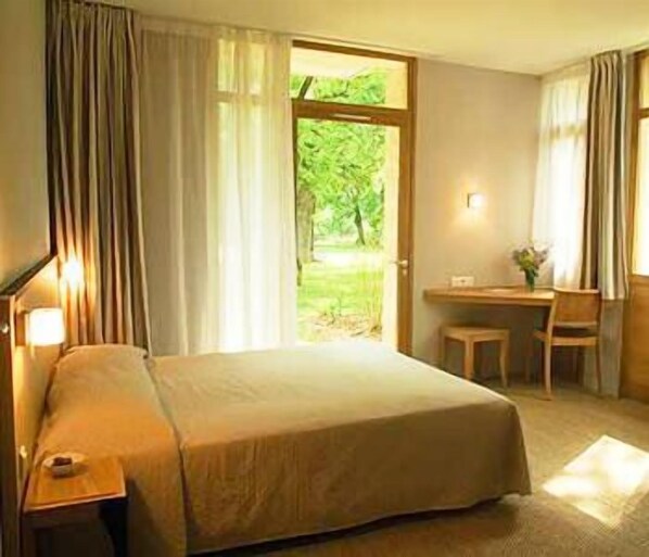 A hotel room with a bed, a desk, and a chair. There is a window with curtains, and a view of greenery outside. - Hôtel Les Esclargies (Rocamadour)