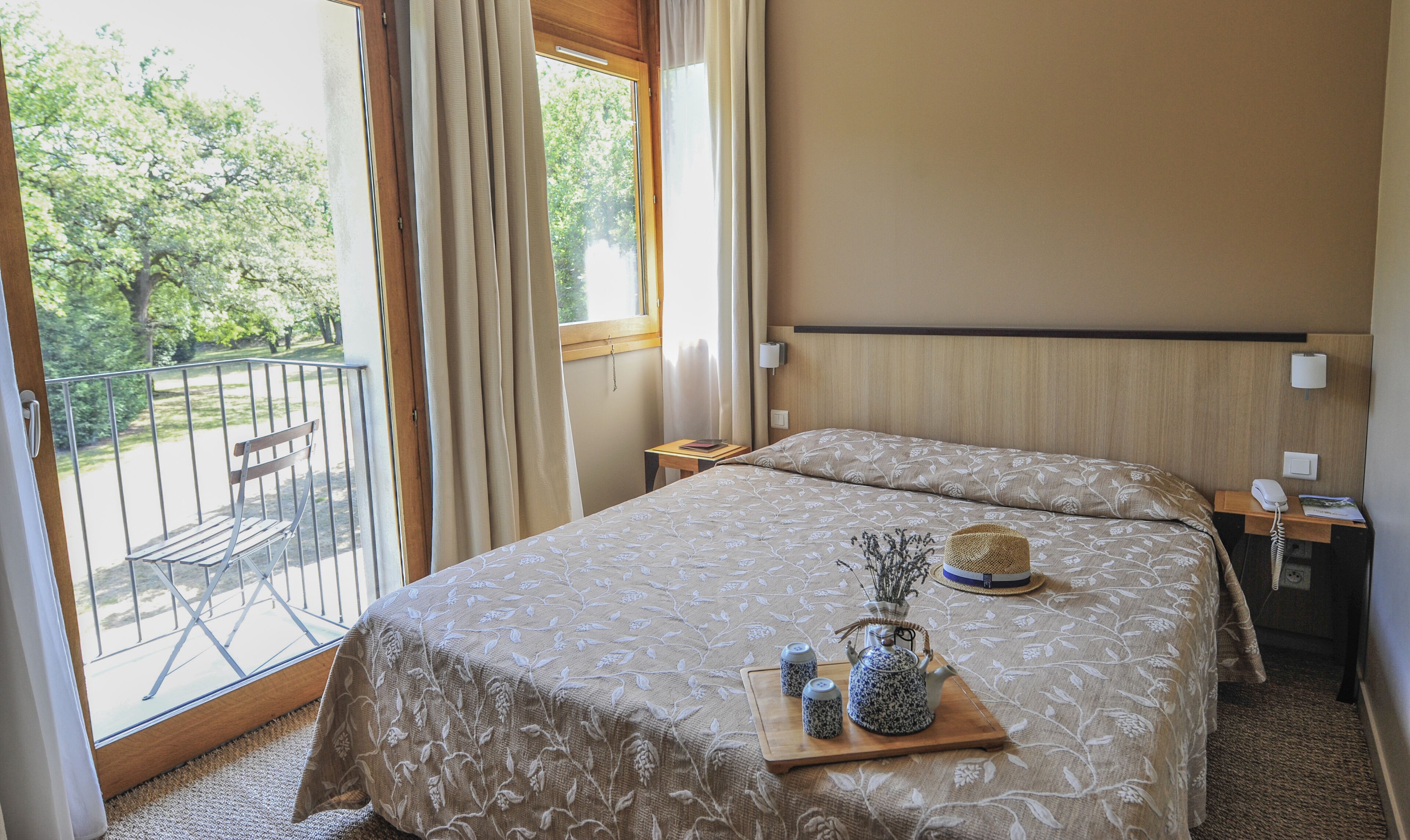 A hotel room with a bed, a bedside table, a nightstand, a window with curtains, a balcony with a chair, and a view of trees.