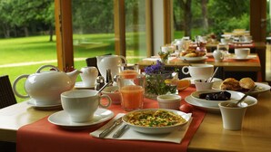 A breakfast table with various dishes, cups, and a teapot, set in a room with a view of a green landscape.