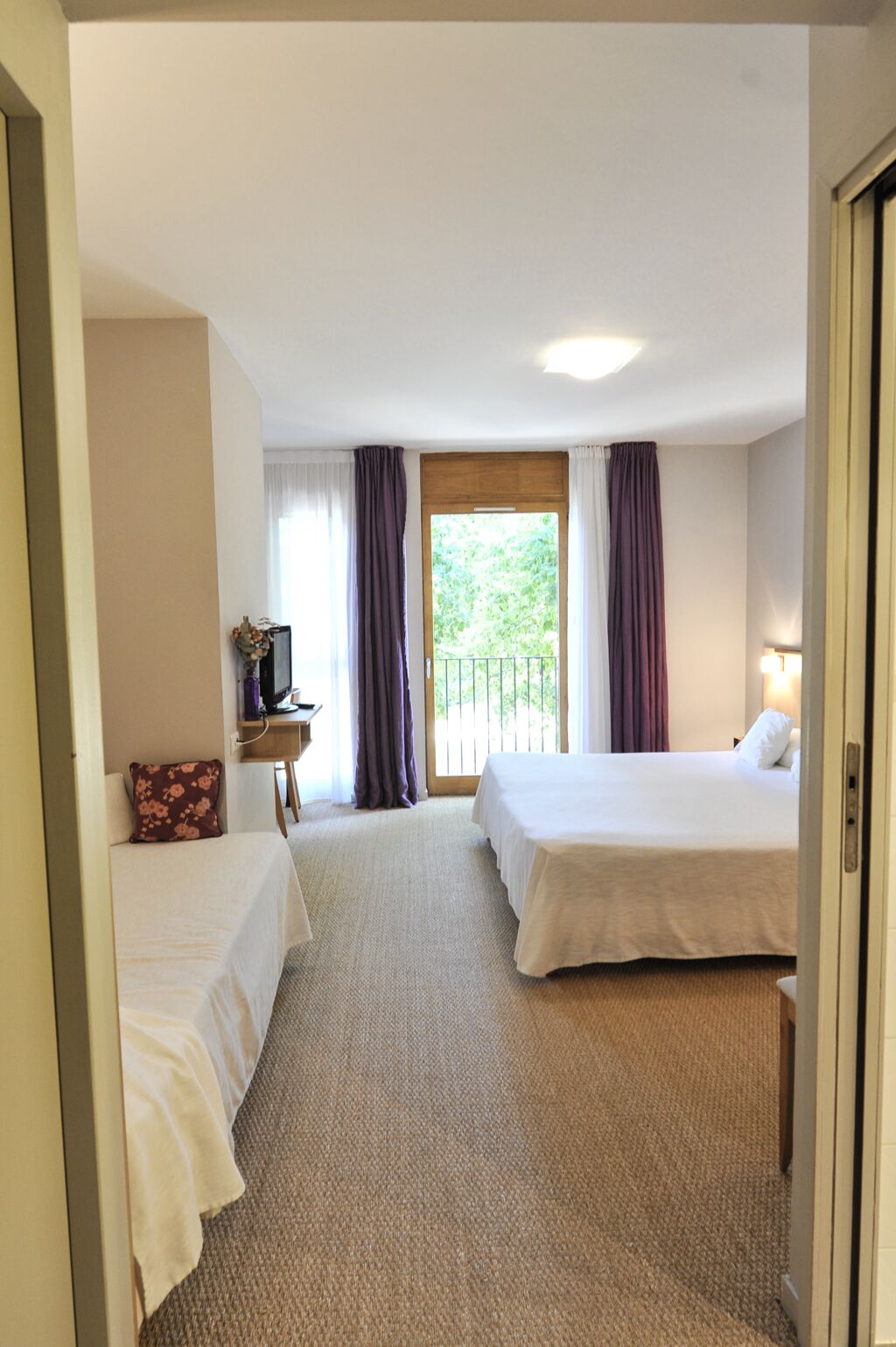 A hotel room with two beds, a small table with a lamp, a window with curtains, and a view of greenery outside.