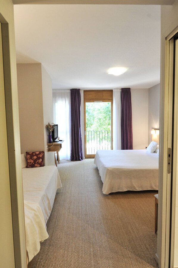 A hotel room with two beds, a small table with a lamp, a window with curtains, and a view of greenery outside. - Hôtel Les Esclargies (Rocamadour)