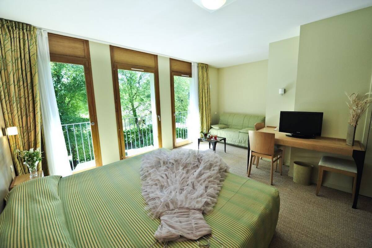 A hotel room with a bed, a desk, a television, and a balcony with a view of trees.