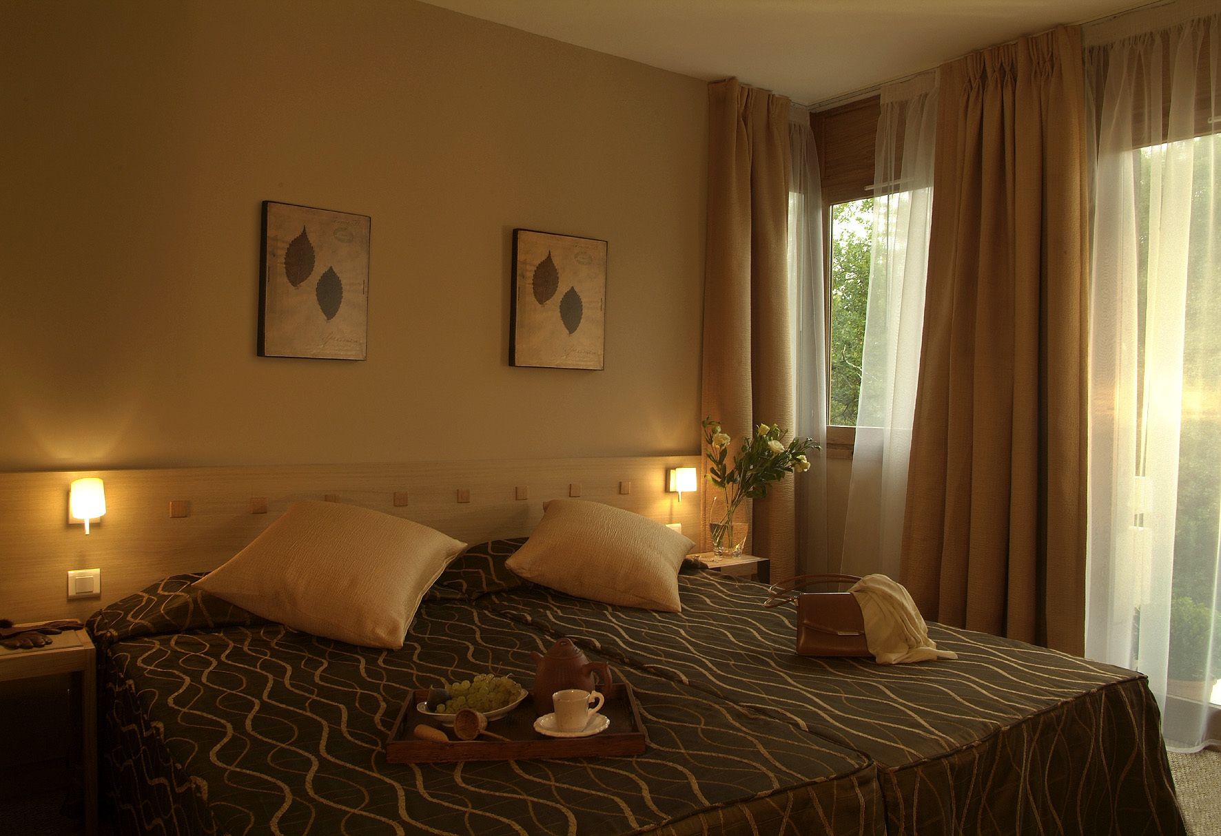 A hotel room with two beds, a bedside table with a lamp, a vase with flowers, and a window with curtains.