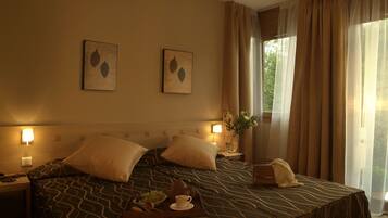 A hotel room with two beds, a bedside table with a lamp, a vase with flowers, and a window with curtains.