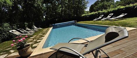 A rectangular swimming pool with a wooden deck, surrounded by lounge chairs and a potted plant.