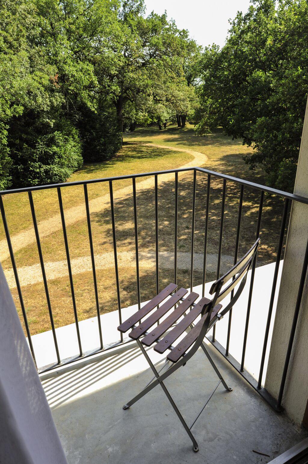 A balcony with a metal railing, a wooden bench, and a view of a park with trees and a path.