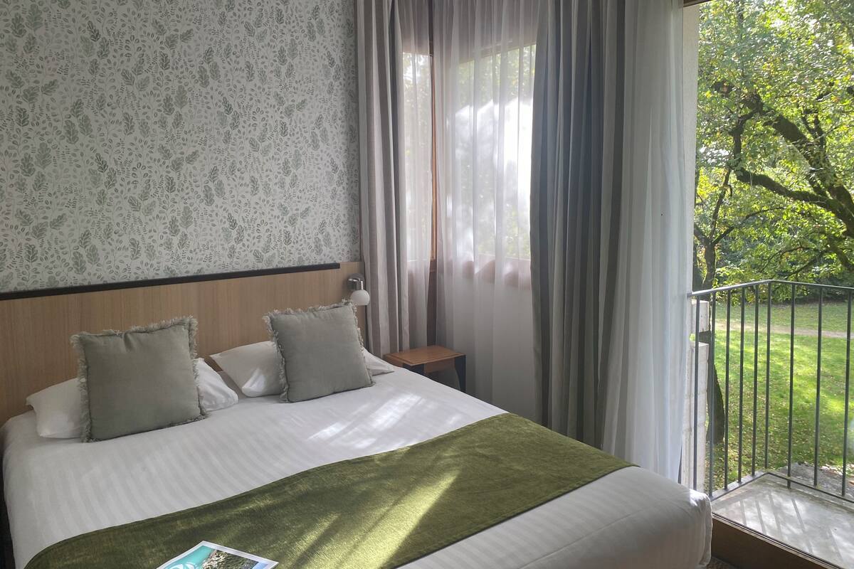 A hotel room with a bed, a nightstand, a lamp, and a view of a green garden through the window.