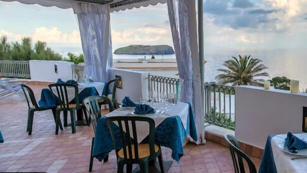 An outdoor dining area with tables and chairs, overlooking a body of water and a distant island. - Calabattaglia (Ventotene)