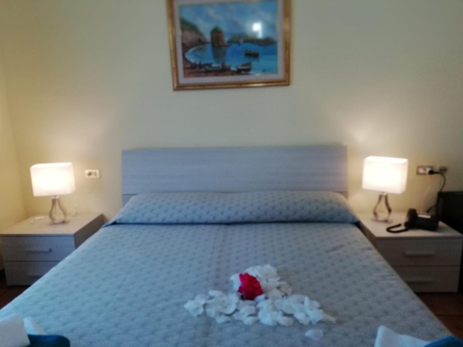 Et hotelværelse med en seng, to sengeborde med lamper, et maleri på væggen og en rød rose på sengen.