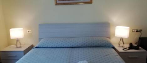 A hotel room with a bed, two bedside tables with lamps, a painting on the wall, and a red rose on the bed.