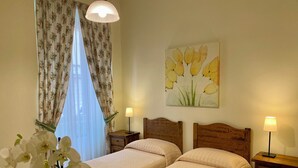 Ein Hotelzimmer mit zwei Betten, einem Nachttisch mit Lampe, einem Holzkopfteil, einem Blumenbild an der Wand und einem Fenster mit floralen Vorhängen.