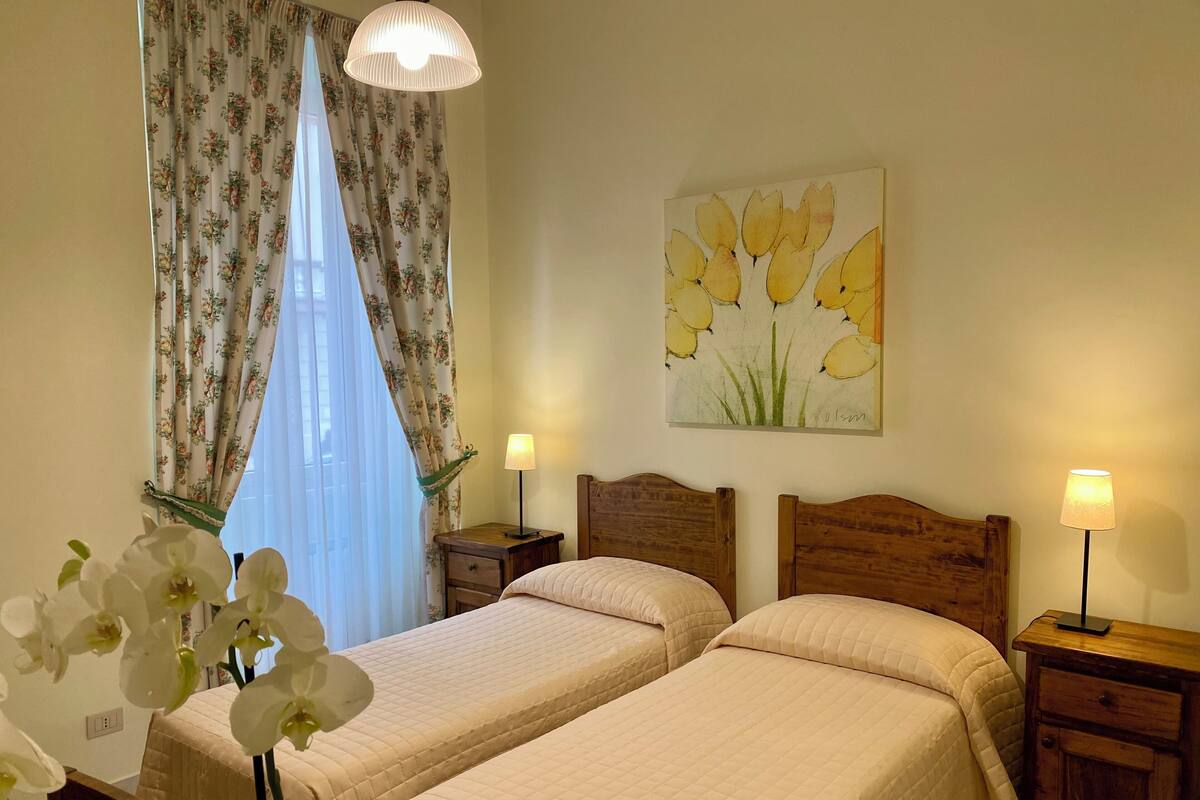 Une chambre d’hôtel avec deux lits, une table de chevet avec une lampe, une tête de lit en bois, un tableau floral accroché au mur et une fenêtre ornée de rideaux à motifs floraux.