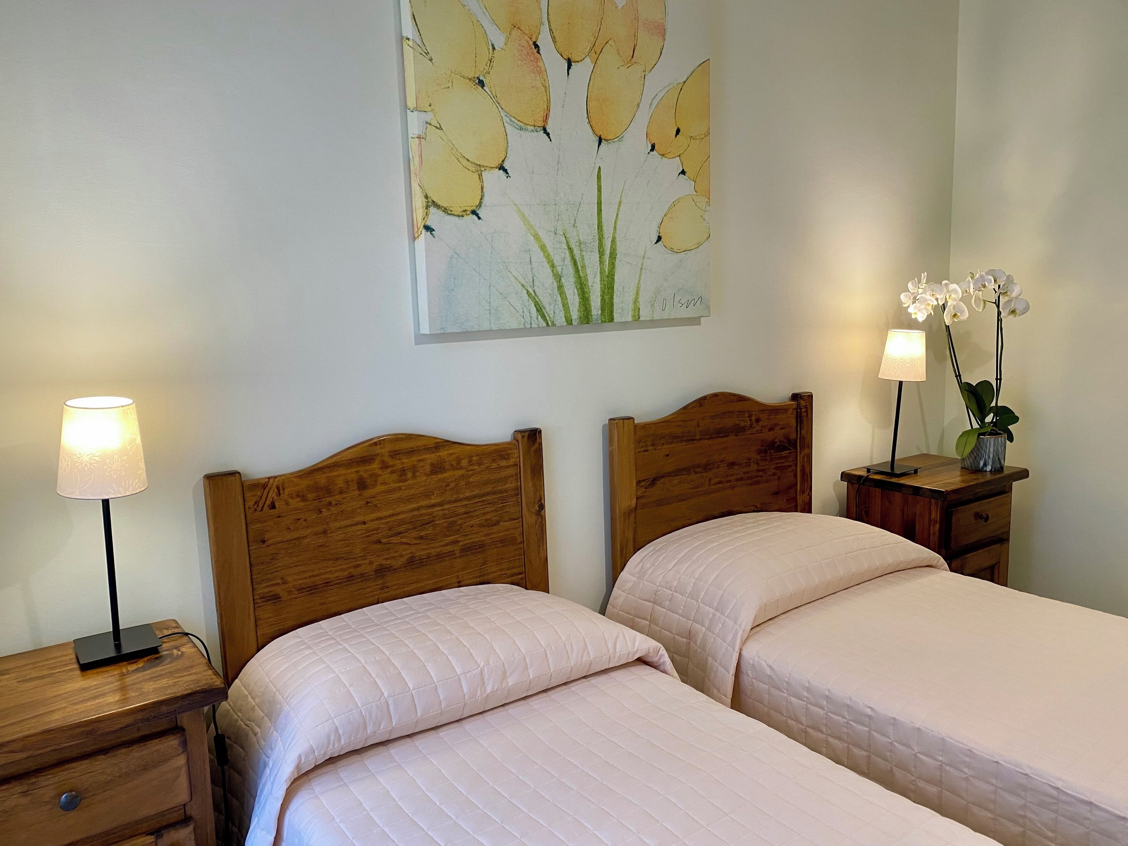 Ein Schlafzimmer mit zwei Holzbetten, einem Nachttisch mit Lampe, einem kleinen Tisch ebenfalls mit Lampe sowie einem Topf mit einer Orchidee.