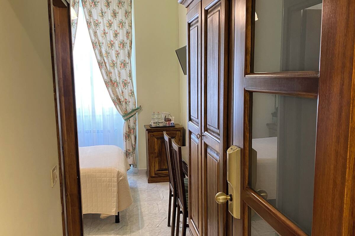 Une chambre à coucher avec un lit, une armoire en bois, une petite table avec une lampe et une fenêtre avec des rideaux.