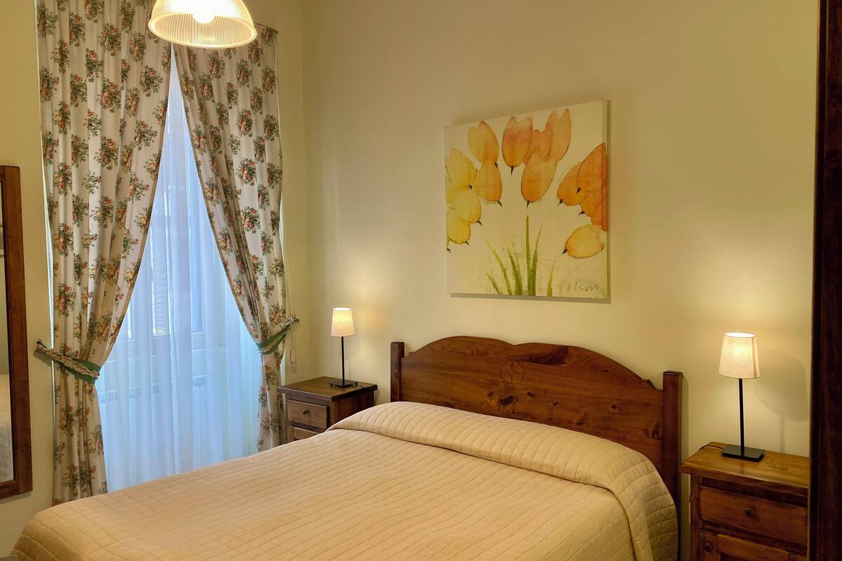 Un lit bien fait, recouvert d’une courtepointe matelassée, avec une tête de lit en bois, deux tables de chevet avec des lampes, un rideau à motifs floraux et un tableau représentant des fleurs accroché au mur.