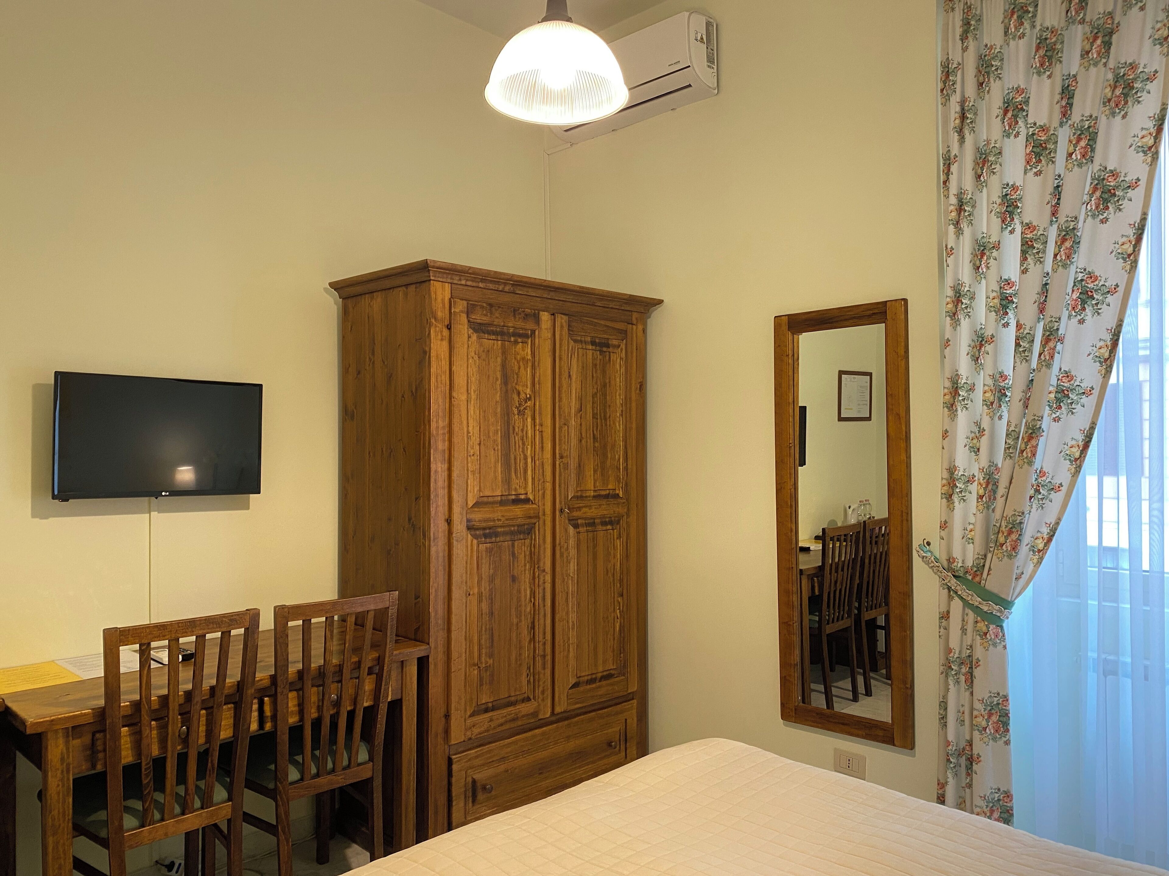 Ein Zimmer mit einem Holzschrank, einem flachbildfernseher an der Wand, einem Schreibtisch mit Stühlen und einem Bett mit gesteppter Matratze.