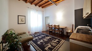 A cozy living room with a wooden ceiling, a dining area with a table and chairs, a kitchen with a sink and countertop, and a large window with curtains. - B&B SubUrbe Roma (Rome)