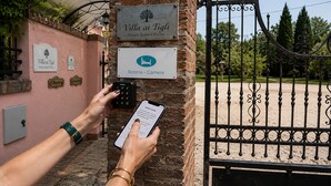 A person using a smartphone to access a door with a keypad, a sign indicating 'Villa ai Tigli - Venice Airport Rooms - Rooms - Camera,' and a gate with a wrought iron design. - Villa ai Tigli Venice Airport Rooms Online Check-in & Keypad Access (Mestre)