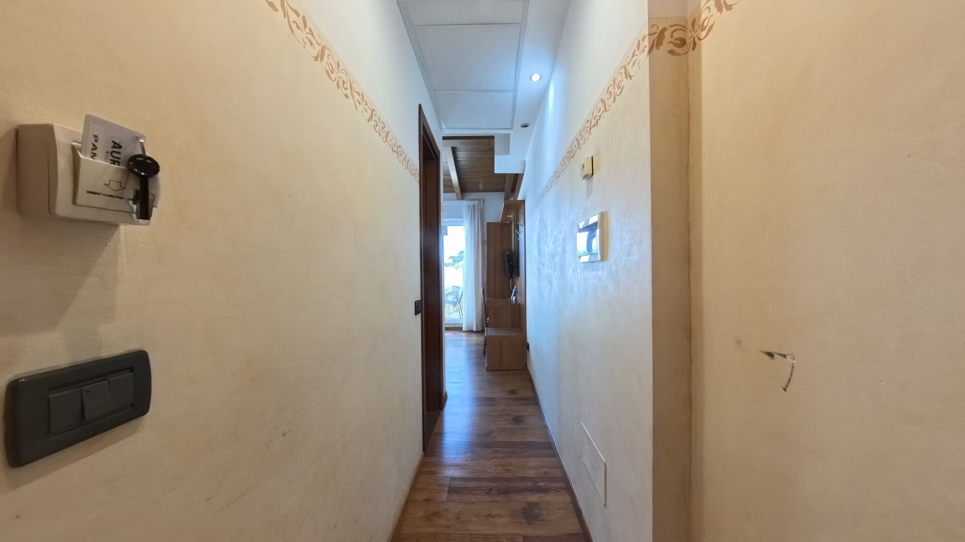 A narrow hallway with a wooden floor, a light switch, and a power outlet on the left wall.