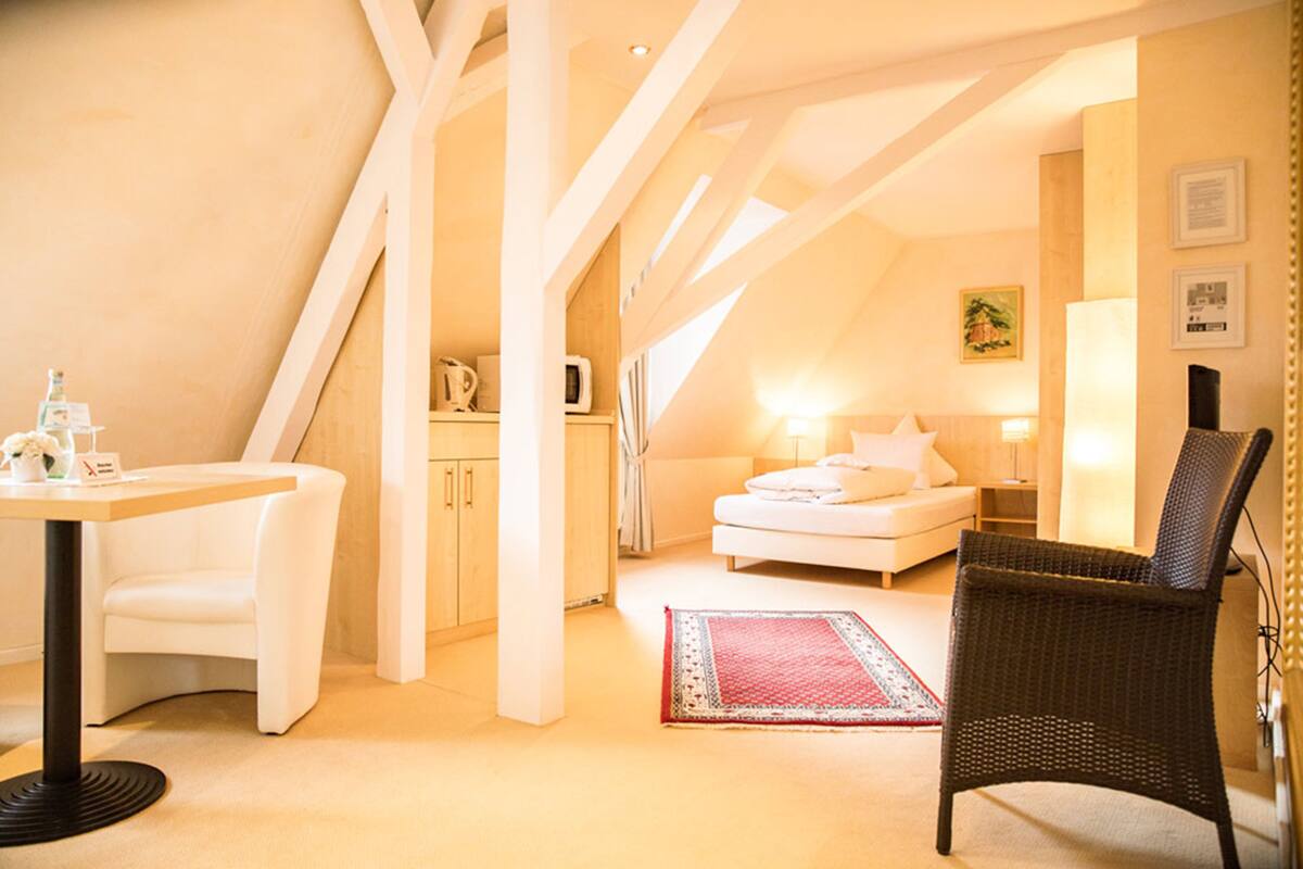 A cozy bedroom with a white bed, a wicker chair, a desk with a chair, and a red rug.