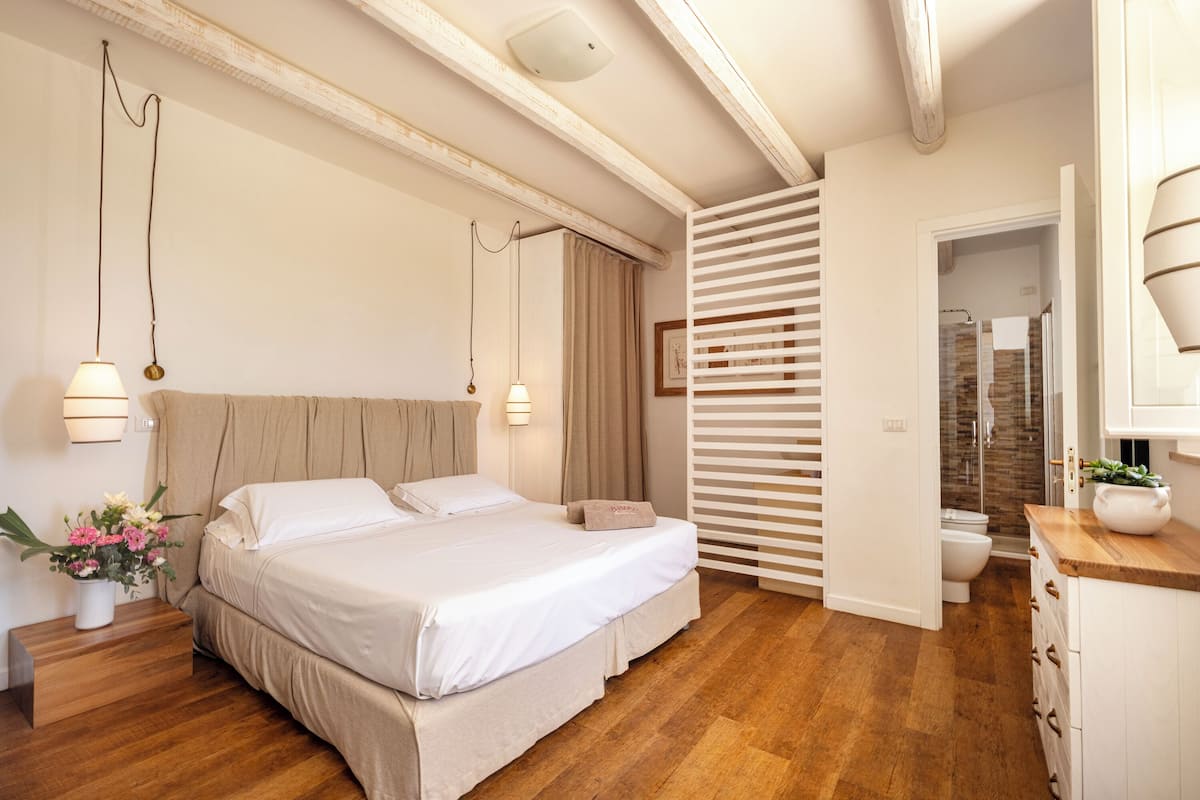 Une chambre avec un grand lit, un plancher en bois, une table de chevet ornée d’un vase de fleurs et une salle de bain visible à travers une porte.