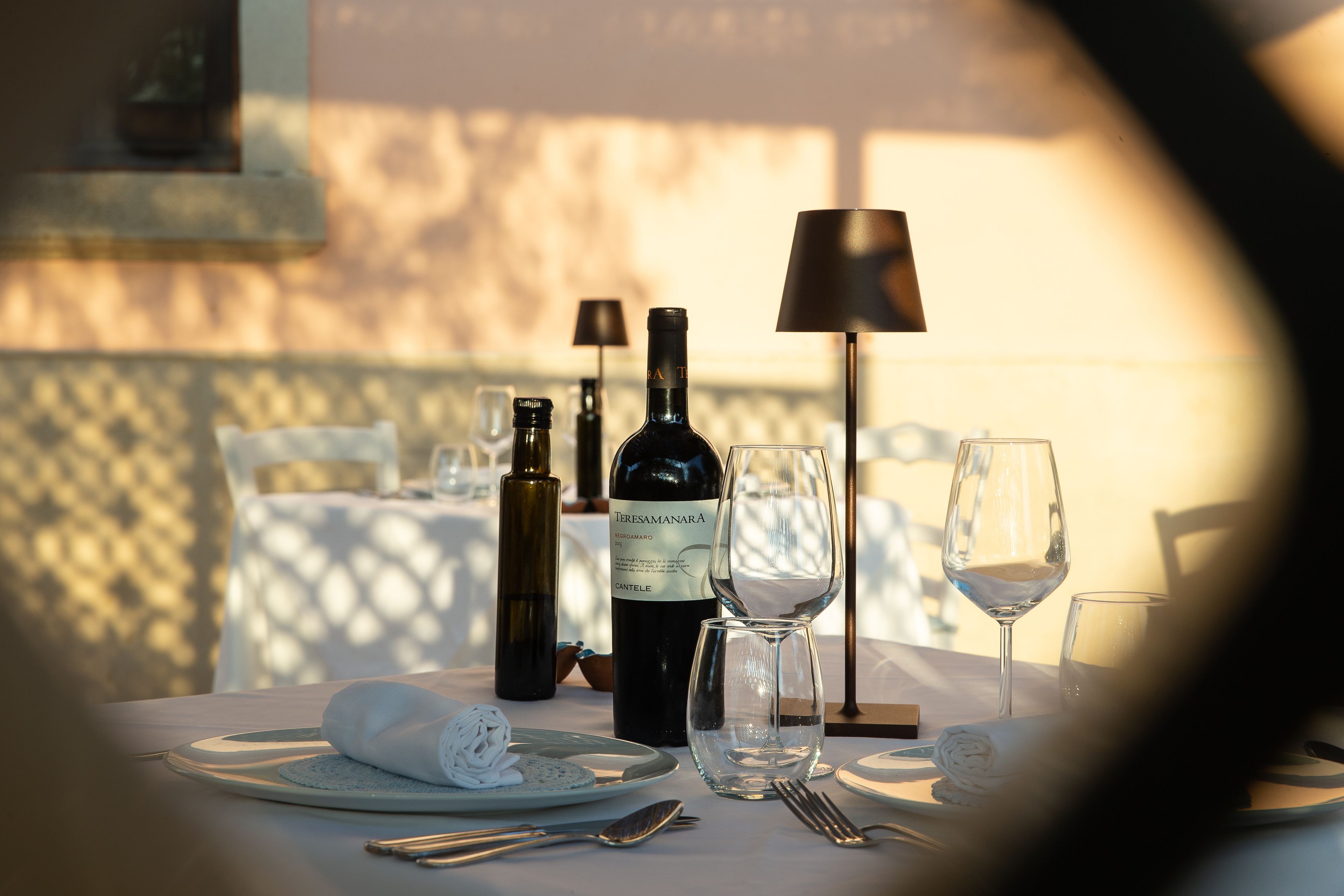 Uma mesa de jantar posta com uma garrafa de vinho, copos e talheres, com uma luminária e uma cadeira ao fundo.