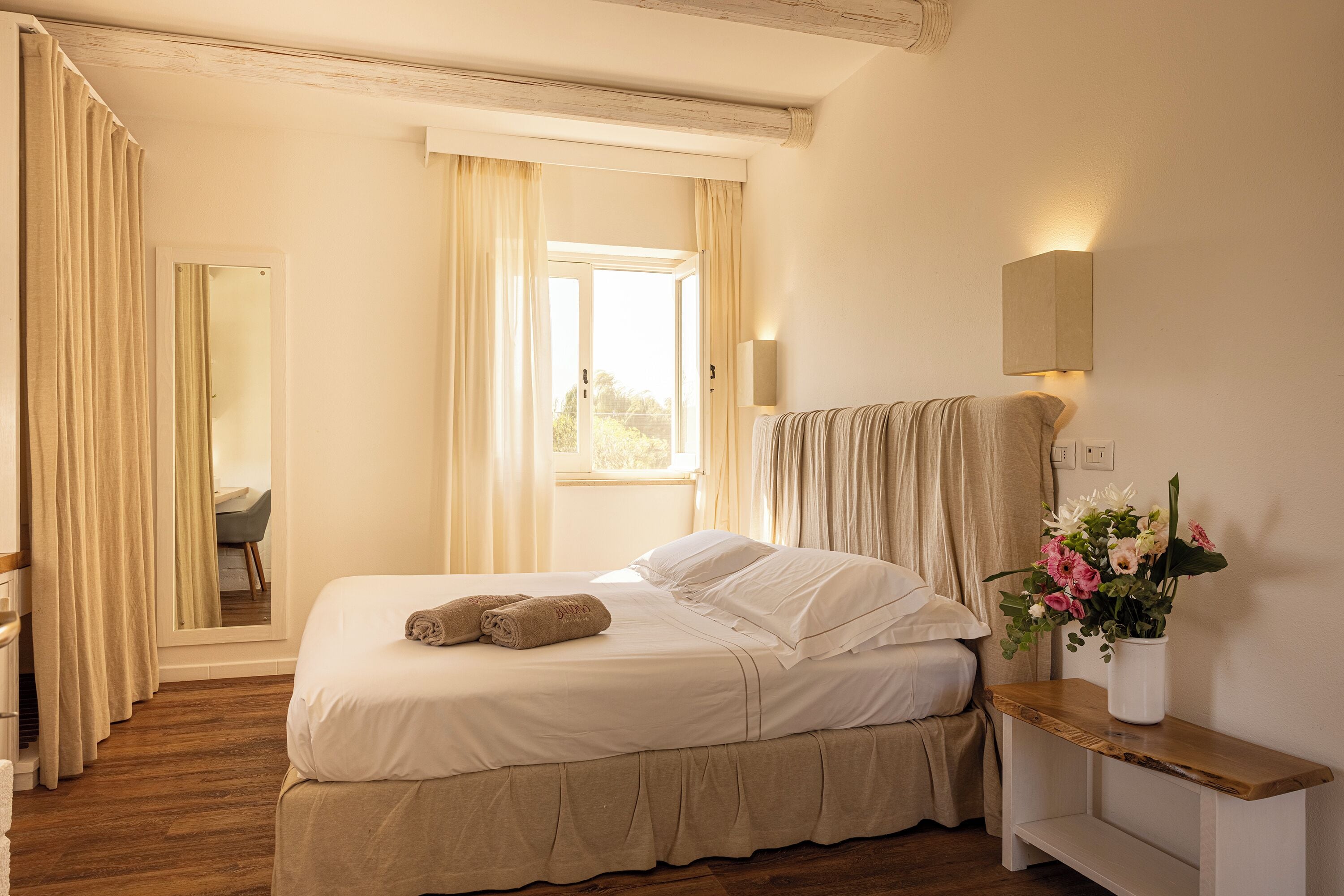 A bedroom with a bed, a bedside table with a vase of flowers, a mirror, and a window with curtains.