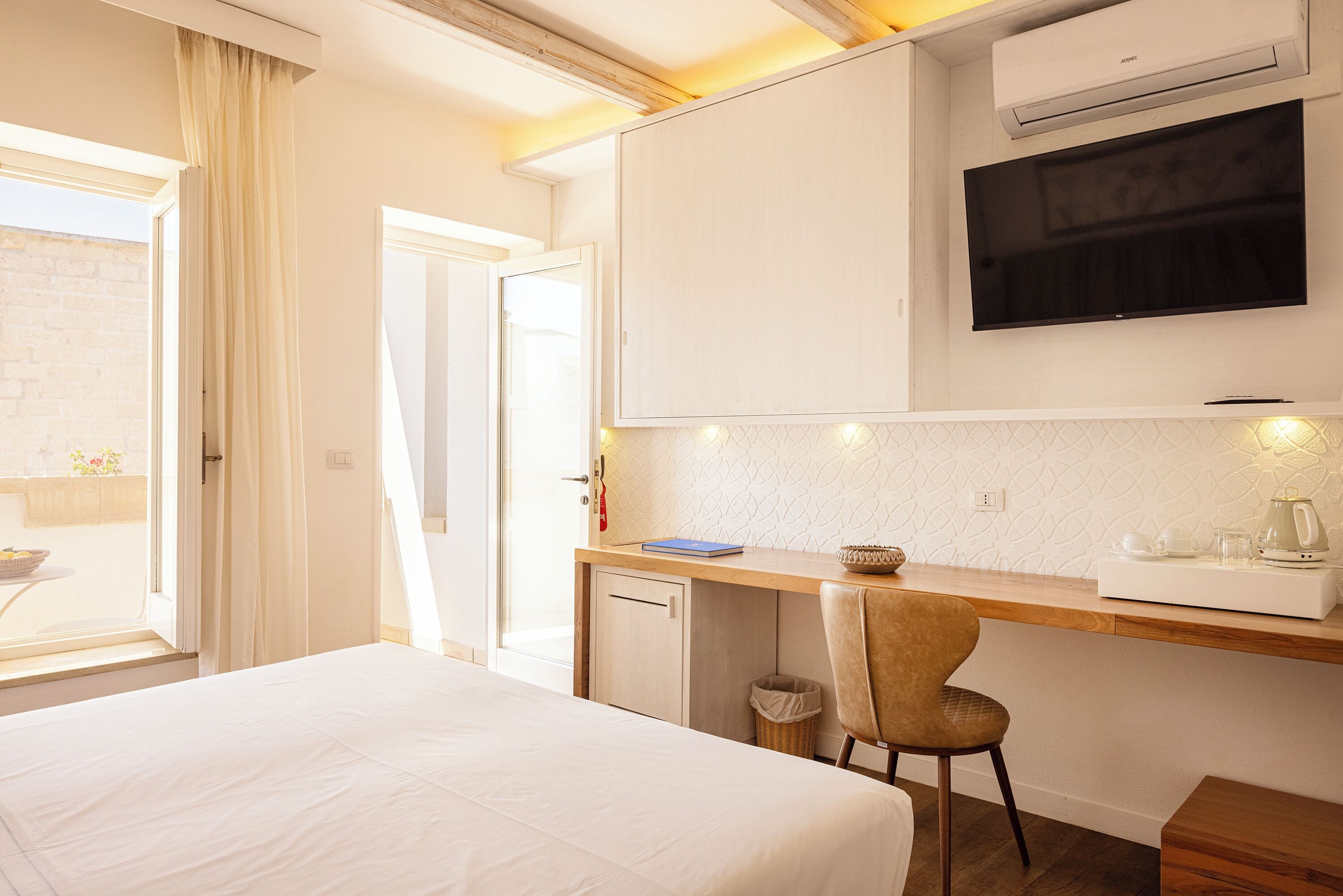 Quarto de hotel com cama, secretária com cadeira, televisão, pequeno espaço de cozinha e janela com cortinas.