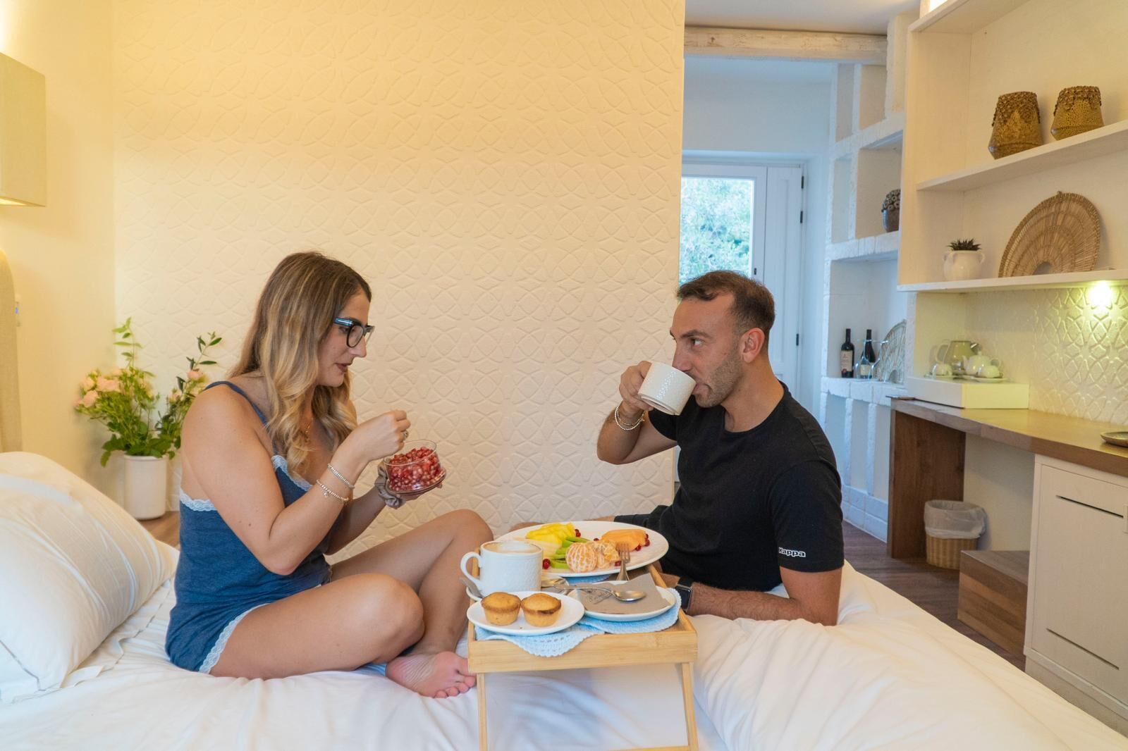 Una pareja sentada en una cama con comida para el desayuno, una mesita y una cocina al fondo.