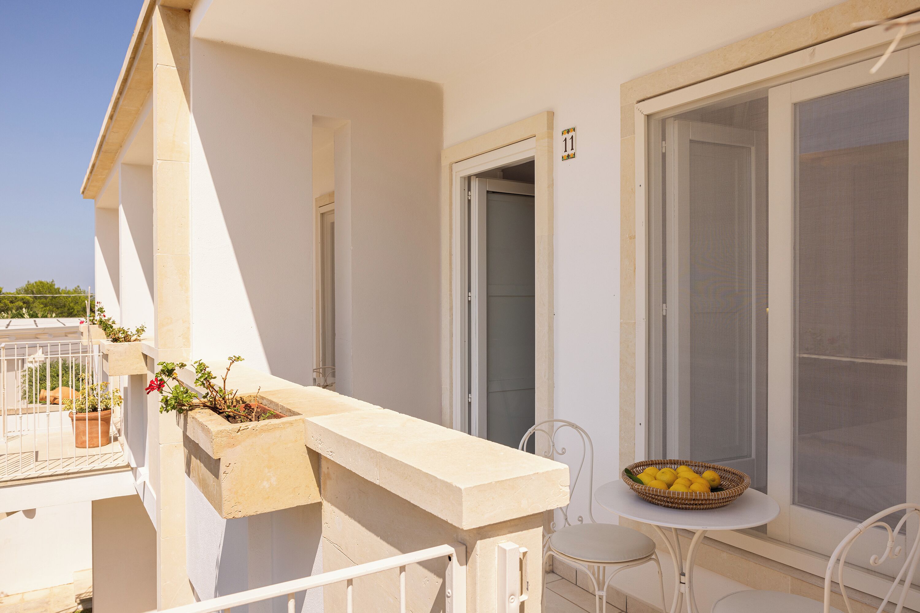 Un balcon avec une balustrade blanche, un pot de fleurs, une petite table sur laquelle se trouve un panier de citrons, et une porte portant le numéro 11.