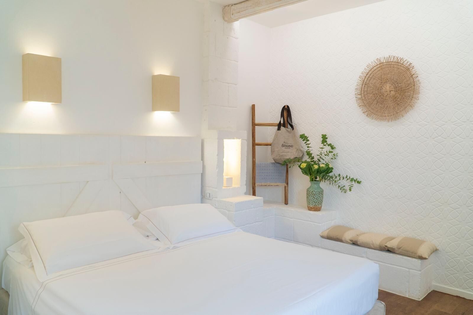 A white bed with pillows, a bedside table, a wall-mounted light, a woven wall decoration, a wooden ladder, a bag, and a vase with flowers.