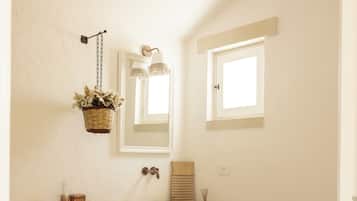Uma casa de banho com toucador bege, lavatório branco, espelho, cesto suspenso com flores e janela.
