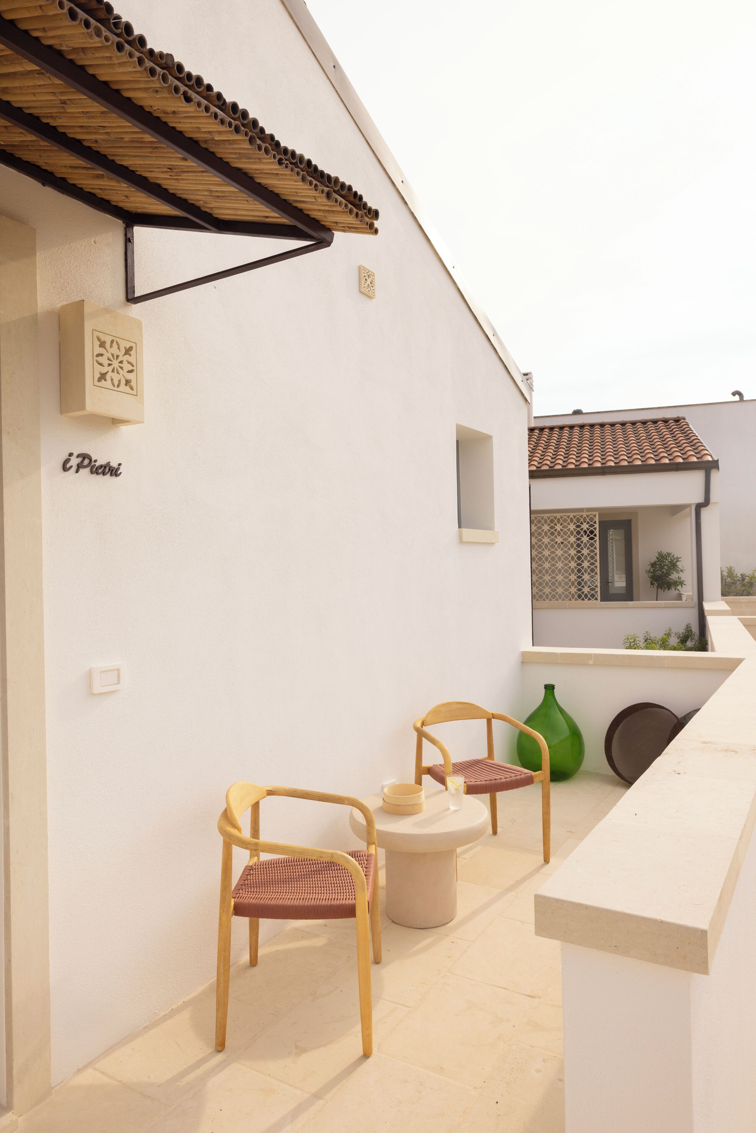 A white exterior wall with a wooden pergola, two wicker chairs, a small round table, and a green bottle.