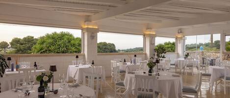 A spacious outdoor dining area with white tables and chairs, set against a backdrop of greenery and a stone wall.