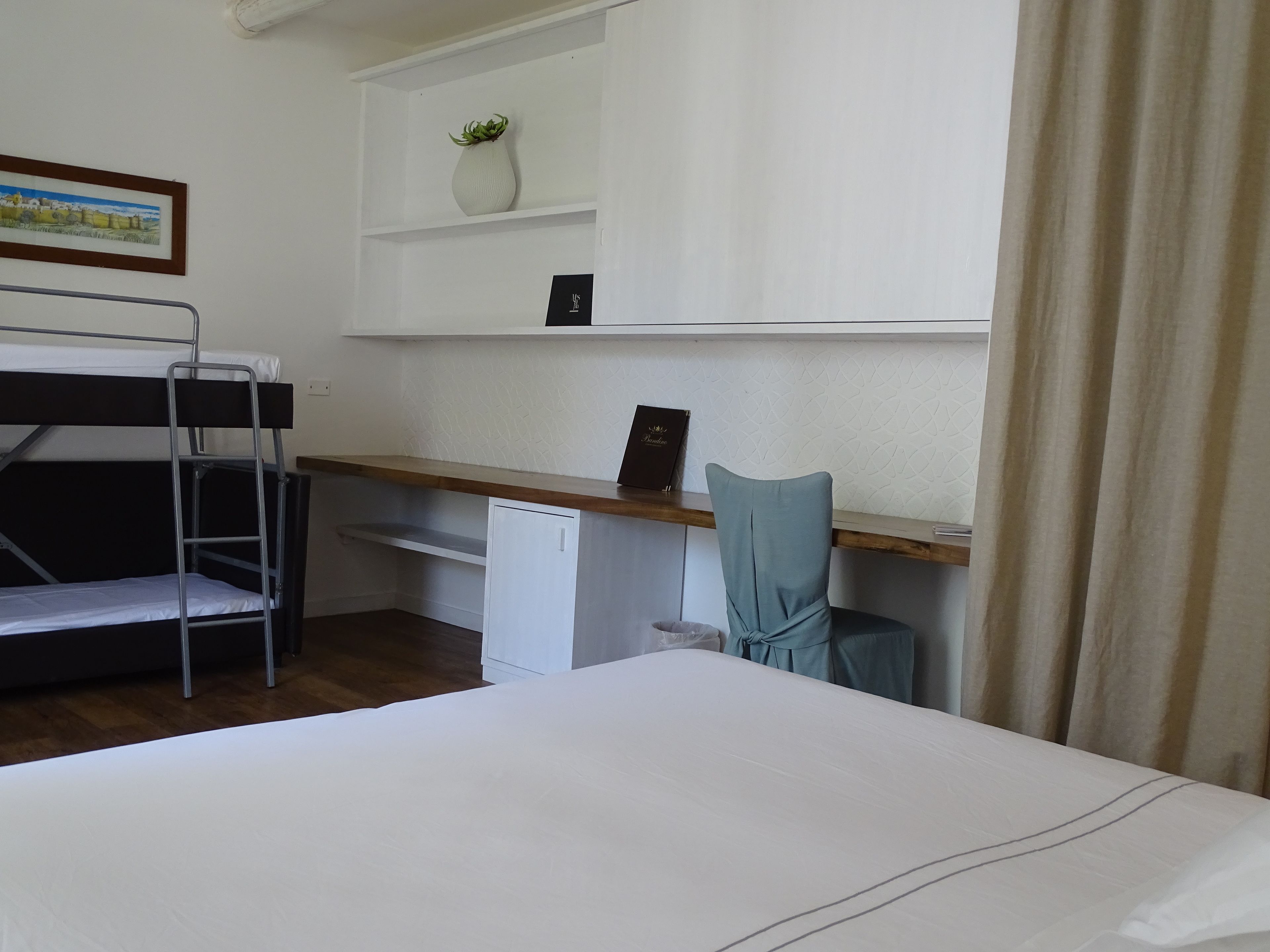 A room with bunk beds, a desk with a chair, a small white cabinet, a framed picture on the wall, and a potted plant on a shelf.