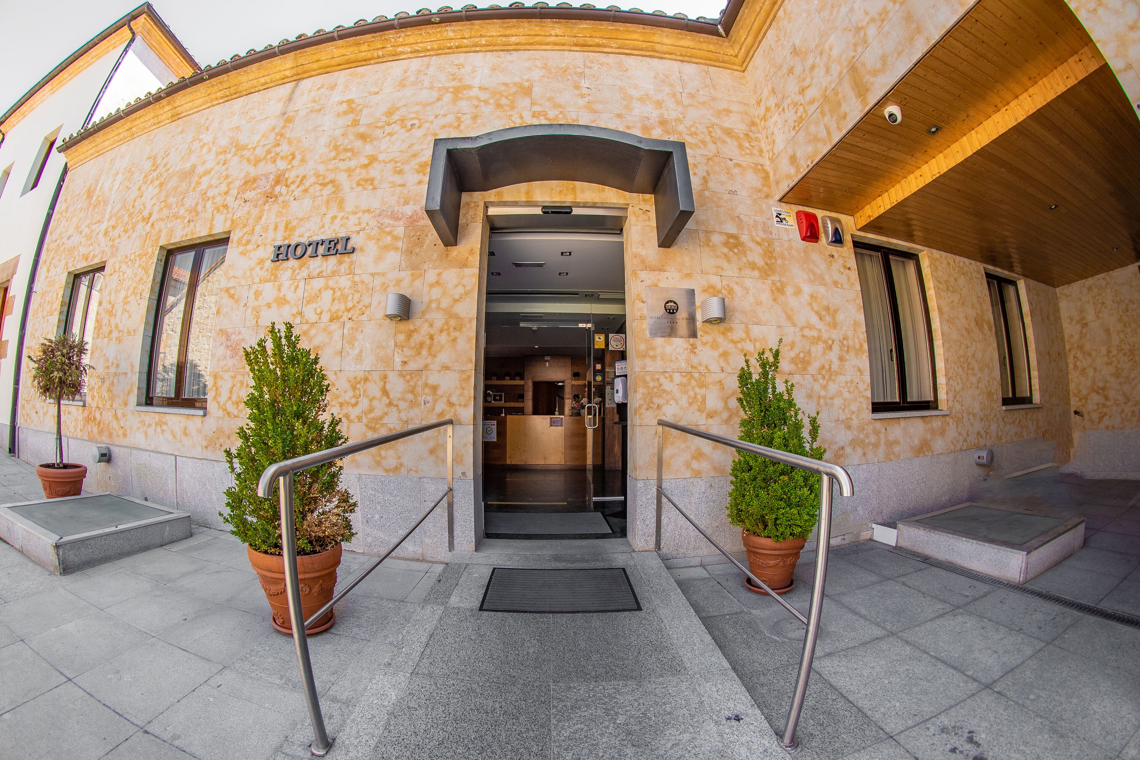 Entrada de hotel con fachada moderna, mostrador de recepción y plantas en macetas.
