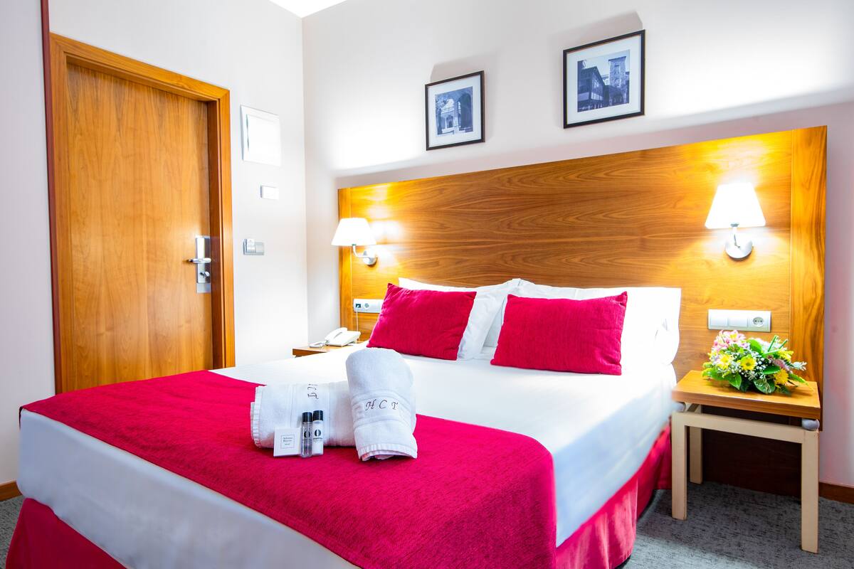Ein Hotelzimmer mit einem hölzernen Kopfteil, einem Bett mit roter und weißer Bettwäsche, einem Nachttisch mit Lampe und einem kleinen Tisch mit Blumenarrangement.