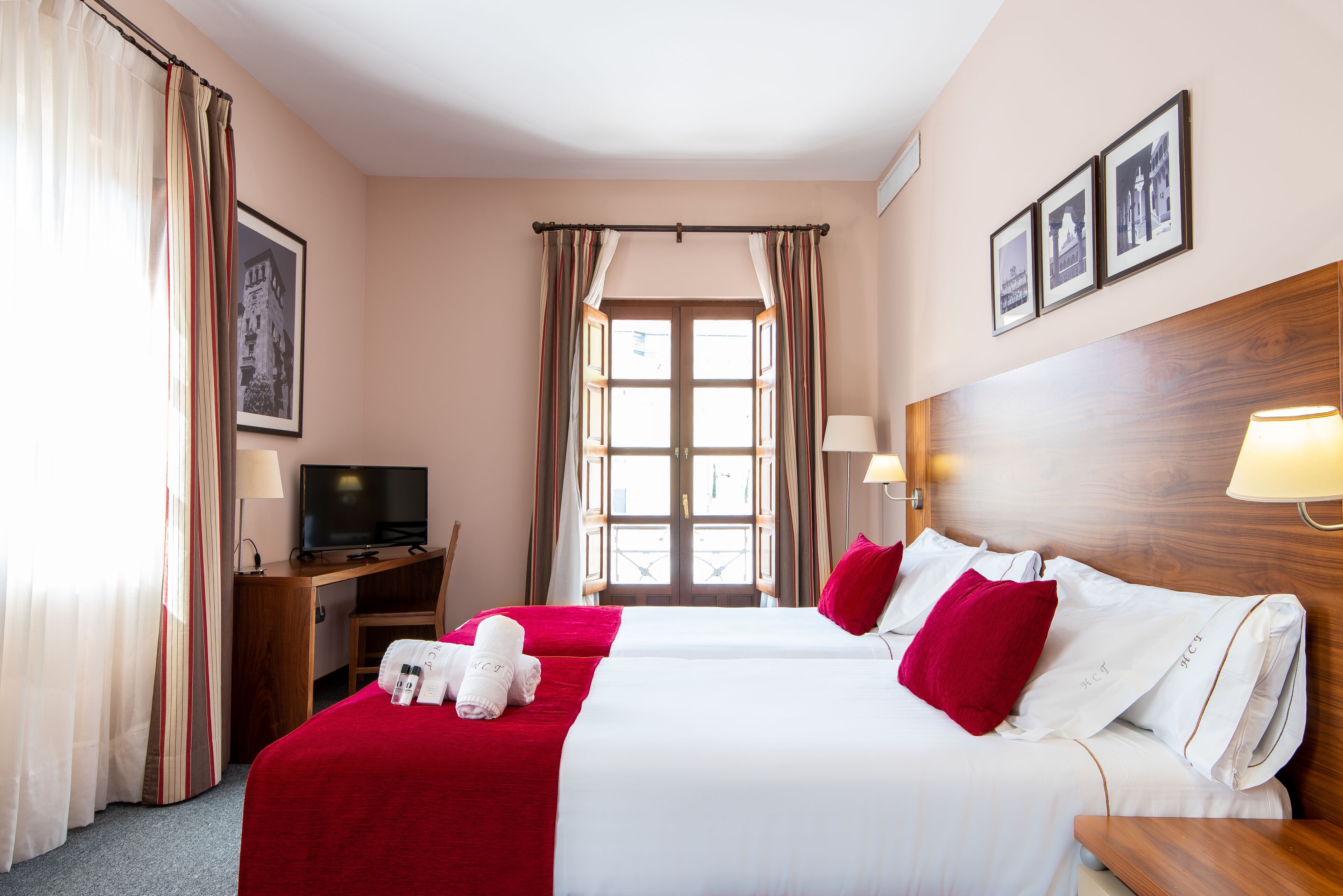 A hotel room with two beds, a desk with a TV, a wooden headboard, and a window with curtains.
