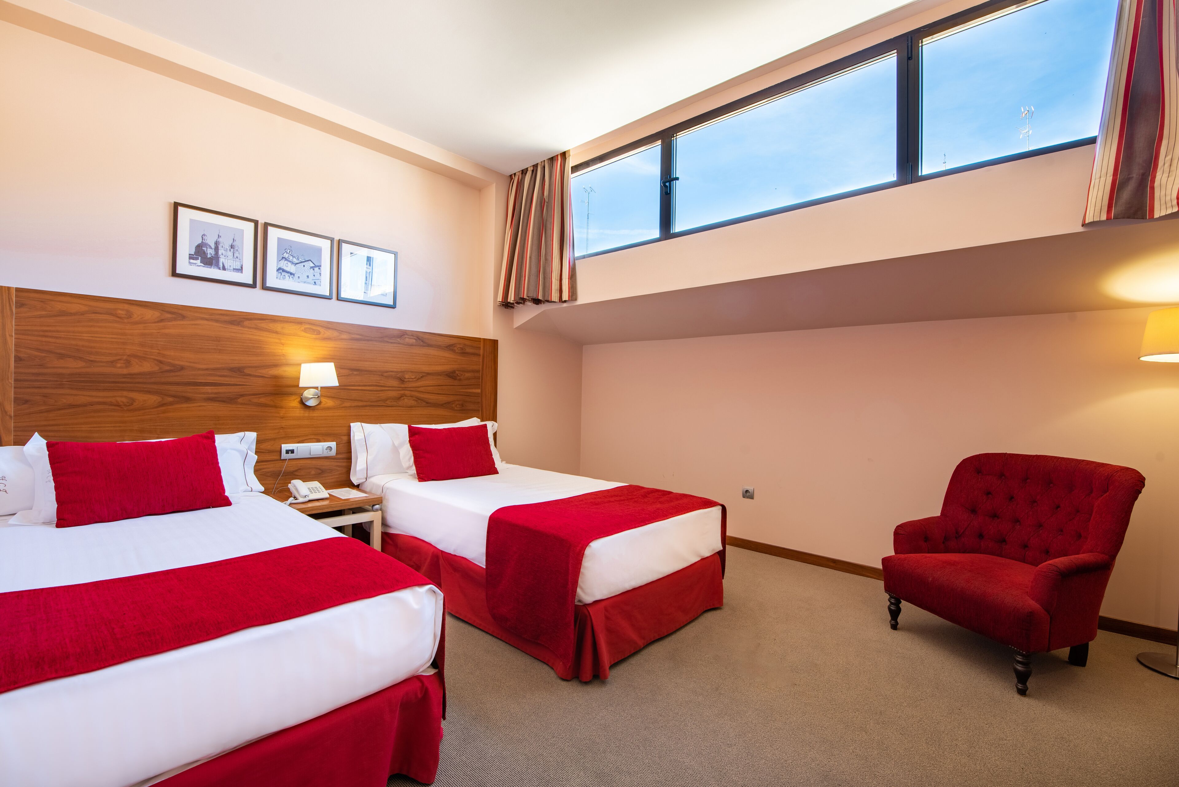 A hotel room with two beds, a red armchair, a wooden headboard, a window with curtains, and wall-mounted artwork.